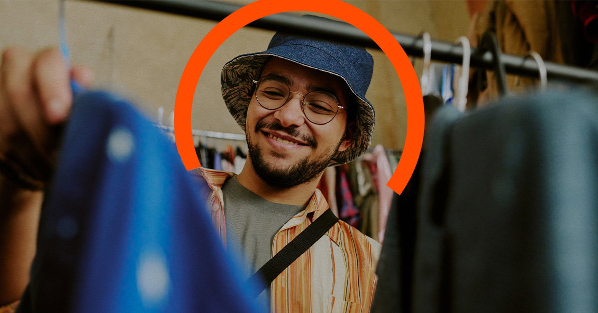 A man wearing a blue bucket hat and glasses smiles while browsing clothes on a rack.